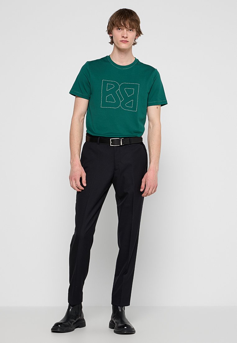 Grünes Baumwoll-T-Shirt mit gesticktem geometrischem Logo, kombiniert mit schwarzen maßgeschneiderten Hosen und schwarzen Knöchelstiefeln, die eine glatte Textur aufweisen.