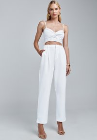 Biały krótki top z skręconym przodem i szerokimi białymi spodniami. Gładka tkanina o minimalistycznym fasonie. Modelka w beżowych sandałach na cienkich paskach.