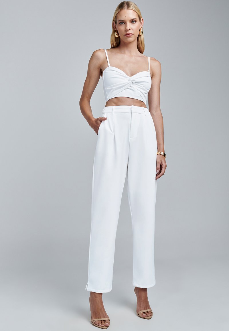 Biały krótki top z skręconym przodem i szerokimi białymi spodniami. Gładka tkanina o minimalistycznym fasonie. Modelka w beżowych sandałach na cienkich paskach.