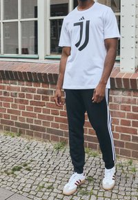 Bílá tričko s černým logem Juventusu, černé tepláky s bílými pruhy a bílé tenisky s červenými akcenty. Pozadí z cihlové zdi.
