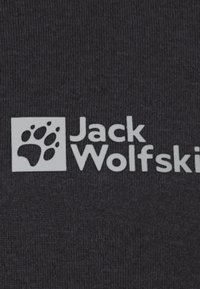 Zwarte stof met een gestructureerde oppervlakte, met een witte logo-box met een pootafdruk en de tekst "Jack Wolfskin" in duidelijke letters.