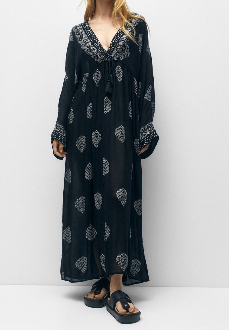 Femme portant une longue robe noire avec des motifs de feuilles blanches et des manches larges, associée à des sandales à plateforme noires sur un fond uni.
