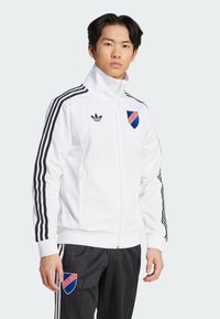 White zip-up athletic jacket made of smooth fabric, featuring black stripes along the sleeves and a blue and red logo on the chest.