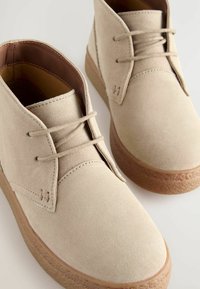 Bottines en suède beige avec un bout arrondi, un laçage à l'avant et une semelle texturée brun. Les coutures minimales et un subtil détail de marque sont visibles.