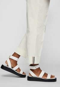 Białe sandały na platformie z regulowanymi paskami wokół kostki, szerokimi paskami na stopę oraz czarną podeszwą, zestawione z kremowymi spodniami z kontrastowym mankietem.