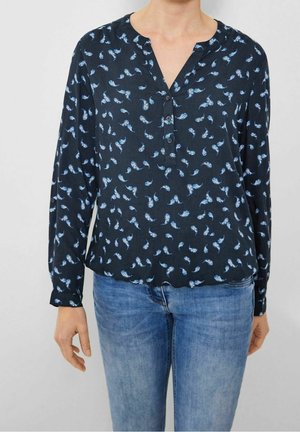 Femme portant un chemisier bleu foncé avec de petits motifs cachemire bleu clair et un jean bleu clair, debout devant un fond blanc uni.
