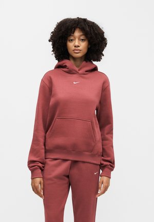 Vrouw met krullend haar, gekleed in een bordeauxrode Nike hoodie en bijpassende joggingbroek, staand tegen een effen witte achtergrond.
