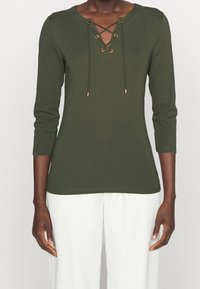 Olivově zelený dlouhý rukáv top s výstřihem do V a šněrováním v přední části; má zlaté kovové kroužky a šňůrky, vyrobený z měkké bavlněné tkaniny.