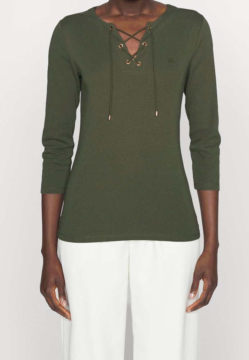 Olivově zelený dlouhý rukáv top s výstřihem do V a šněrováním v přední části; má zlaté kovové kroužky a šňůrky, vyrobený z měkké bavlněné tkaniny.