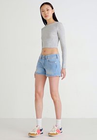 Un haut cropped à manches longues gris associé à un short en denim bleu clair. Le modèle porte des baskets multicolores et des chaussettes blanches mi-mollet. Fond simple.