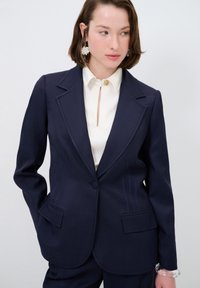 Blazer sartorial blu navy con chiusura a un bottone, due tasche laterali e dettagli di cucitura sottili. Abbinato a una blusa di colore chiaro.