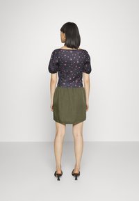 Top preto de flores com mangas curtas e design justo, combinado com uma saia verde, franzida na cintura, que fica acima do joelho, e sandálias de salto.