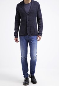 Mörkblå stickad kavaj med krage, knappfront och två fickor, i kombination med ljusblå jeans och svarta skor.