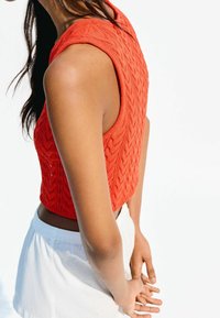 Rood gebreid croptop met een getextureerd chevronpatroon, mouwloos ontwerp en een afgeronde halslijn, gecombineerd met witte korte broek.