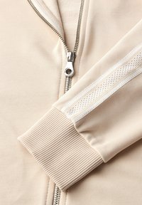 Primo piano di un indumento beige con zip, dotato di cerniera metallica e polsini a coste, decorato da una striscia bianca con dettagli in rete.
