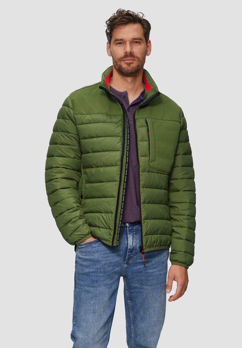 Grüne Puffjacke mit Reißverschluss, ausgestattet mit einem hohen Kragen, einer Brusttasche und einem kontrastierenden roten Innenfutter. Getragen über einem lila Hemd.