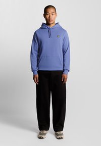 Hættebluse i lys blå bomuld med en frontlomme og broderet logo. Sammen med sorte bukser og grå sneakers.