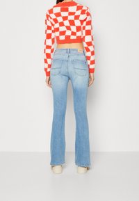 Kvinna i ljusblå utsvängda jeans, krämfärgade platåskor och en röd-vit rutmönstrad kort sweater, stående vänd bort.