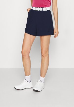 Woman wearing navy blue shorts with white belt, white socks, and white athletic shoes, standing against a plain light background.