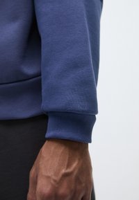 Sudadera de color azul marino con puños acanalados, textura suave y diseño ajustado. Se puede ver parcialmente una mano agarrando el puño.