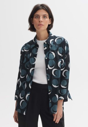 Camicia nera con motivi circolari bianchi e blu, maniche lunghe, chiusura a bottoni e una vestibilità comoda, indossata sopra una maglietta bianca e pantaloni neri.