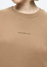 Μπεζ φούτερ κατασκευασμένο από απαλό ύφασμα με στρογγυλή λαιμόκοψη. Διαθέτει ένα διακριτικό λογότυπο κεντημένο σε γκρίζο χρώμα στο μπροστινό μέρος.