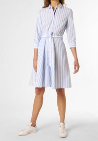 Kvinna som bär en knälång blå och vit randig skjortklänning med knytband i midjan och vita sneakers, stående mot vit bakgrund.