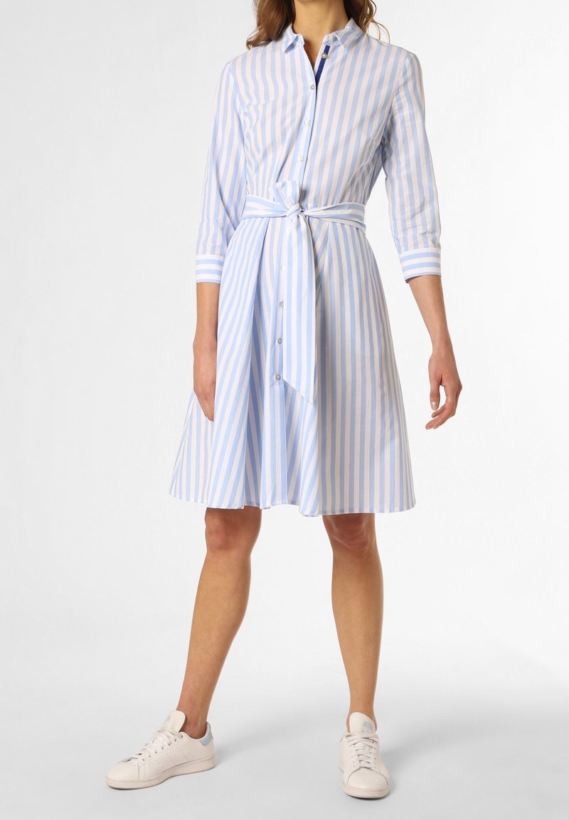 Kvinna som bär en knälång blå och vit randig skjortklänning med knytband i midjan och vita sneakers, stående mot vit bakgrund.