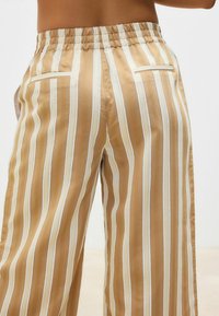 Striped trousers in light brown and cream, made of smooth fabric. Features include an elastic waistband and back pockets.