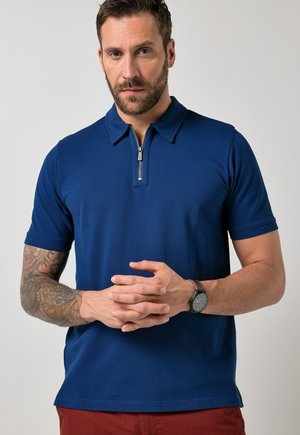 Homme avec barbe et bras tatoué portant un polo bleu à manches courtes avec fermeture éclair, une montre-bracelet et un pantalon rouge, debout devant un fond uni.