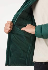 Personne portant une veste verte montre la main à l'intérieur de la poche intérieure, vêtue d'une chemise blanche et d'un pantalon noir, sur un fond uni.