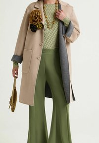 Cappotto beige in misto lana con accenti grigi, abbinato a una maglia verde con fantasia e pantaloni verdi a zampa. Completo di spilla floreale e una piccola borsa dorata.