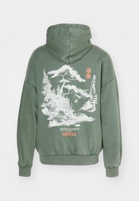 Vihreä huppari, jossa valkoinen havupuiden ja vuorten graafinen kuva selässä sekä oranssit tekstit ja yksityiskohdat alareunassa.