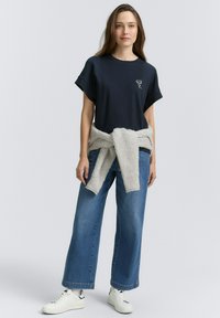 T-shirt bleu marine à manches courtes avec un design brodé, associé à un jean large bleu et un pull en tricot gris clair noué à la taille. Baskets blanches.