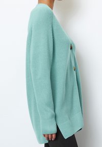 Mint green knit cardigan featuring a relaxed fit, long sleeves, and wooden buttons. Textured fabric with ribbed cuffs and hem.