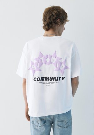 Mladý muž stojí chrbtom, má na sebe biele oversize tričko s fialovými hviezdami a nápisom "COMMUNITY" na chrbte.