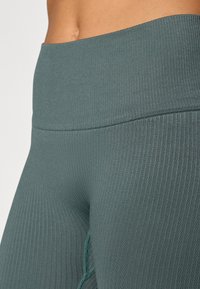 Leggings em teal com textura estriada e cintura alta, apresentando uma textura suave e costuras em contraste; a vista de perto destaca o material e o design.