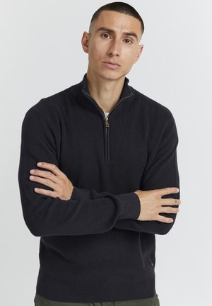 Sweater noir à col zippé en texture côtelée, doté d'une petite fermeture éclair et de poignets ajustés. Confectionné en tissu doux et chaud.