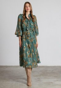 Vestido azul verdoso con patrón floral dorado, botones acampanados y mangas abullonadas. Combinado con botas de ante beige hasta la rodilla. Tejido ligero y texturizado.
