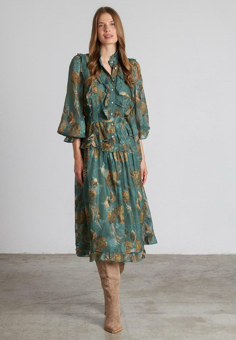 Vestido azul verdoso con patrón floral dorado, botones acampanados y mangas abullonadas. Combinado con botas de ante beige hasta la rodilla. Tejido ligero y texturizado.