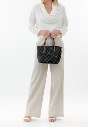 Woman in cream blouse, wide-leg trousers, and white sandals holding a black quilted handbag with brown handles in front of her.