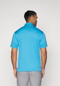 Camisa polo azul con mangas cortas, tela texturizada, cuello y logo de Under Armour en la parte posterior. Se lleva con pantalones de color gris claro.