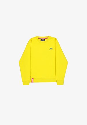 Jasnożółta bluza z długim rękawem i okrągłym dekoltem, z małym niebieskim logo "Alpha Industries" na piersi oraz czerwoną metką materiałową przy krawędzi.