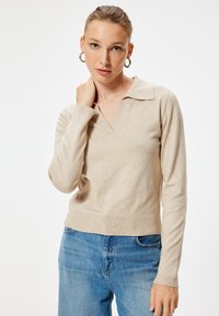 Beżowy sweter dzianinowy z dekoltem w serek i prążkowanym wykończeniem. Długie rękawy, miękka tekstura, w zestawieniu z jasnoniebieskimi dżinsami.