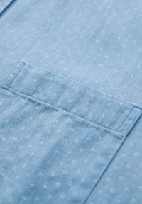 Lichtblauwe stof met kleine witte geometrische patronen, voorzien van een borstzak met zichtbare stiksels en een textuurrijke oppervlakte.