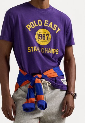 Man draagt een paarse T-shirt met opdruk "Polo East 1967 State Champs", een blauw-oranje gestreepte trui om zijn middel gebonden en grijze joggingbroek.