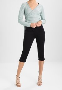 Jasnozielona bluzka wrap z głębokim dekoltem w serek, połączona z czarnymi spodniami 3/4. Modelka nosi taupe sandały na obcasie z paskami, stojąc na neutralnym tle.