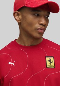 Ung mand iført en rød kasket og rød Puma sweatshirt med Ferrari-logo og hvide buede stikninger på en grå baggrund.