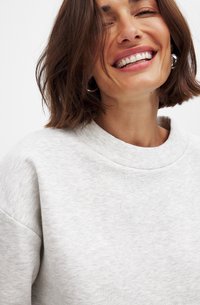 Lichtgrijze sweatshirt met een ribgebreide ronde hals en een zachte textuur. Dichtbij opname van de stofdetails en een glimlachend gezicht met halflang bruin haar.
