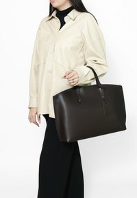 Sac fourre-tout en cuir marron avec une forme structurée, des poignées supérieures et un petit détail de tag ; associé à une chemise oversize beige clair.